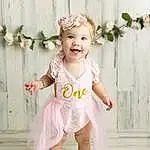 Clothing, Face, Smile, Dress, Baby & Toddler Clothing, Sleeve, Happy, Iris, Pink, Baby, Toddler, Day Dress, One-piece Garment, Barefoot, Fun, Peach, Embellishment, Waist, Child, Headpiece, Person, Joy, Headwear